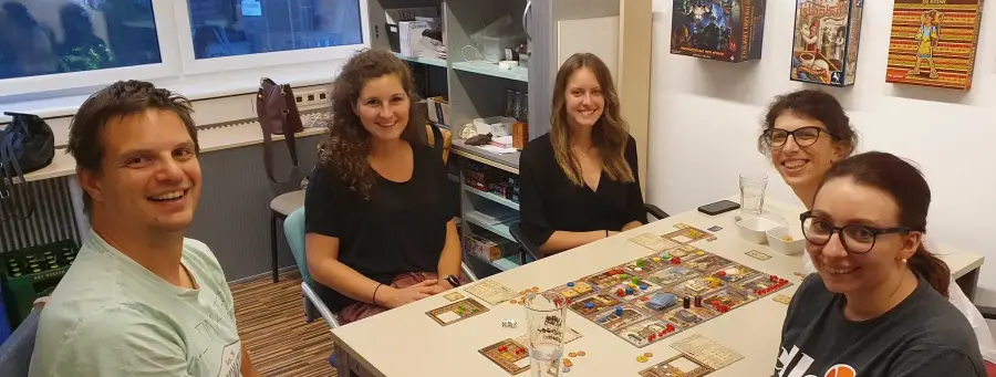 Auf dem Foto sitzen vier Frauen und ein Mann um einen Spieltisch, auf welchem ein Brettspiel aufgebaut ist. Alle fünf Personen schauen in die Kamera und Lachen dabei. Im Hintergrund befindet sich ein Regal mit Spielutensilien und es wurden die Deckel von ausgewählten Spielen an der Wand aufgehängt.